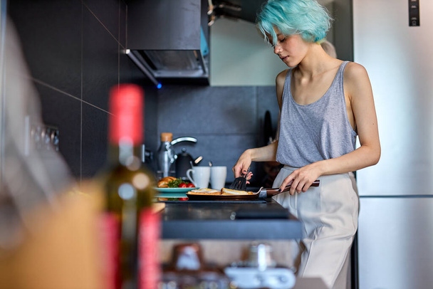 Attractive young woman with modern green hairstyle is cooking breakfast, side view - Valokuva, kuva