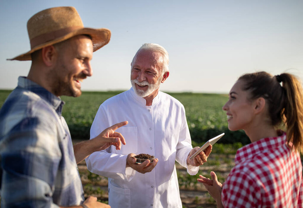 畑に立つ土の質について話し合うカップルの若い農民笑顔。タブレットとペトリ皿を用いた実験用コートを着た高齢者. - 写真・画像