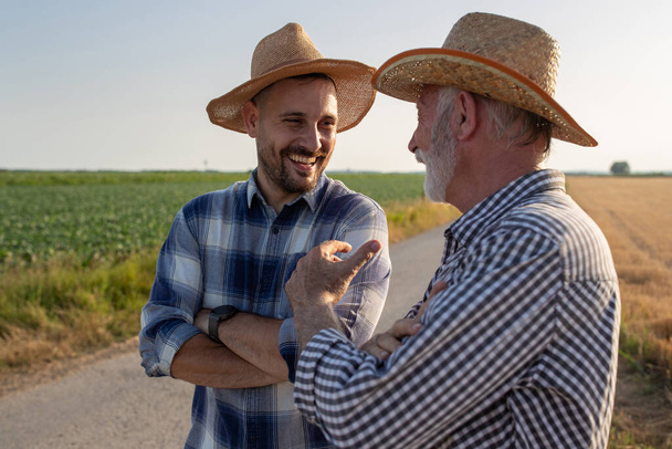 Young and elderly farmers standing on road next to field with crossed arms. Two men wearing plaid shirts and straw hats talking laughing.  - Fotoğraf, Görsel