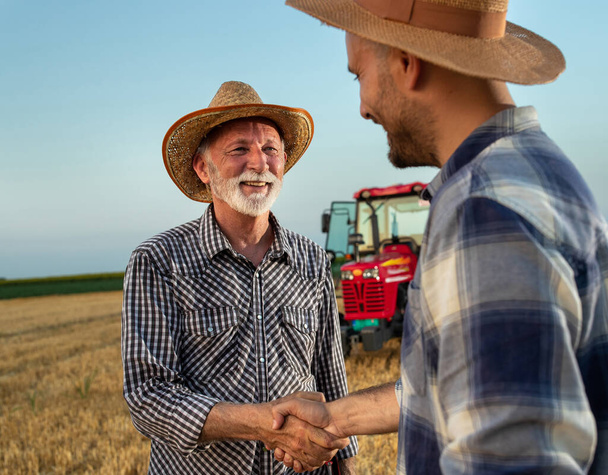 пожилой фермер пожимает руку молодому фермеру, стоящему на собранном поле с трактором на заднем плане. - Фото, изображение
