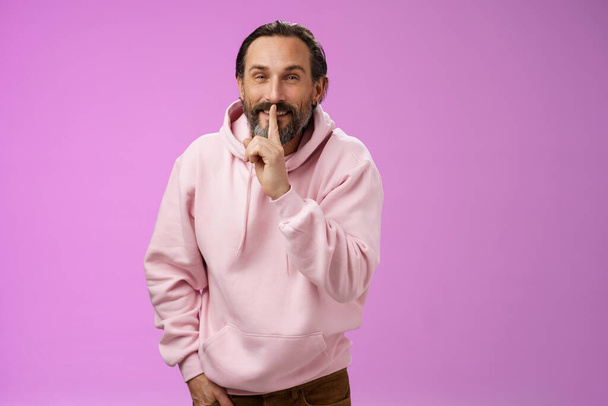 Lips sealed secret. Portrait alluring cheeky stylish adult bearded guy in pink hoodie asking keep quiet show shush shh gesture index finger pressed mouth smiling devious smug face, have idea - Photo, Image