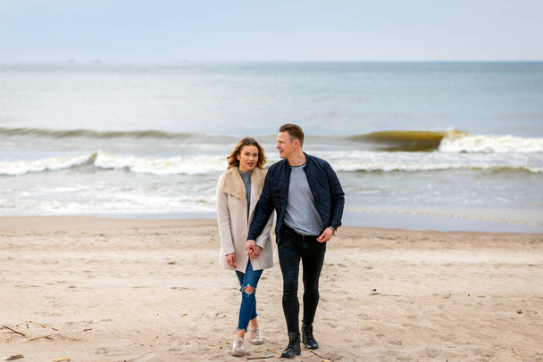 Attractive young couple walking along the shore of a sandy beach, on a spring romantic holiday, outdoors. Travel tourists lifestyle. Couple enjoying loving time together. - Photo, Image