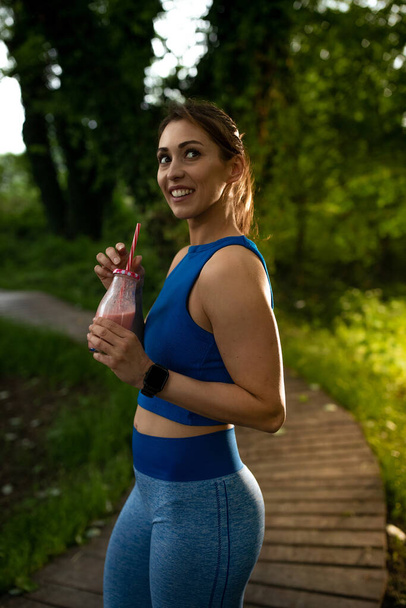 Vrouwelijke atleet houdt smoothie kijken naar kant glimlachen. Meisje nemen pauze na het sporten met milkshake in de natuur.  - Foto, afbeelding