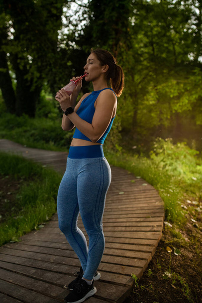 Jovem atleta hidratante descansando após o exercício. Mulher fazendo pausa bebendo smoothie usando palha no caminho na natureza.  - Foto, Imagem