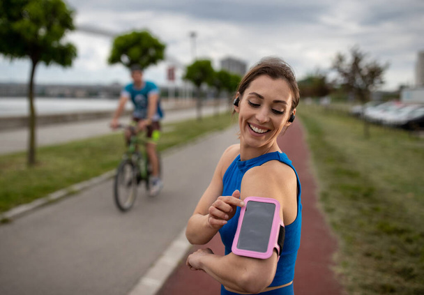 Portrait of young athlete preparing to jog outdoors in urban area. Young woman standing in front of cyclist setting up music on smartphone before run.  - Foto, immagini