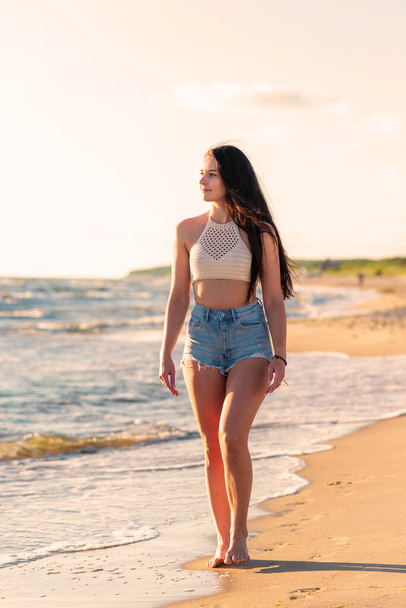 Sahilde gün batımında yürüyen yalnız genç bir kadın. Plaj kadını yaz tatilinde okyanus suyunun yanında rahatça yürüyor.. - Fotoğraf, Görsel