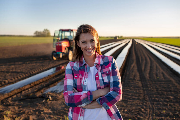 Женщина-фермер, стоящая со скрещенными руками, смотрит в камеру. молодой фермер, стоящий в поле с растительной фольгой и улыбающимся трактором. - Фото, изображение