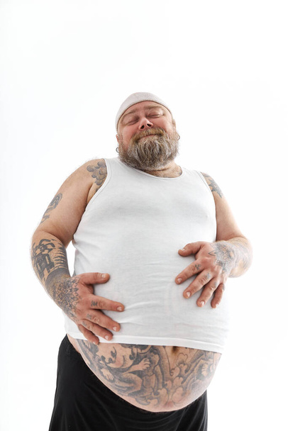 Onnellinen lihava mies, jolla on iso vatsa ja tatuoinnit urheiluvaatteissa, pitää vatsaansa. - Valokuva, kuva