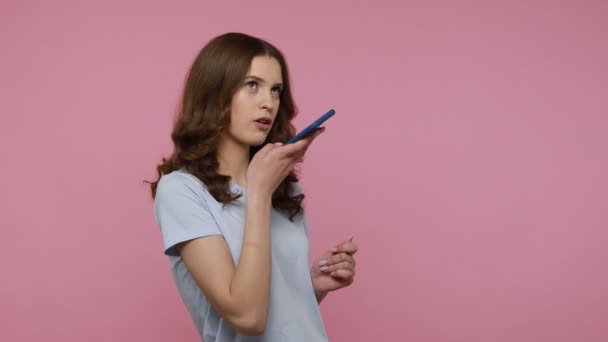 Attractive woman in blue T-shirt making voice records on mobile phone, talking to smart device, using virtual assistant for online search. Indoor studio shot isolated over pink background. - Metraje, vídeo