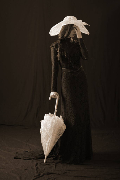 A girl in an old dress and hat stands on a dark background and is sad - Foto, afbeelding
