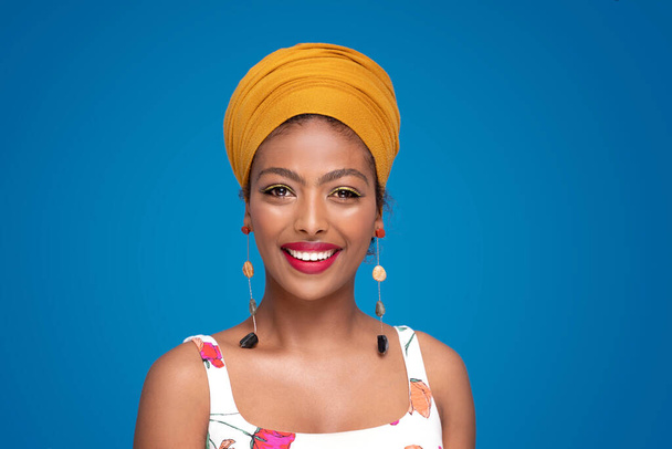 Beautiful happy afro american ethnic girl with turban on her head smiling to the camera, posing on blue pastel studio background. Culture. Real people emotions. Fashion. Colorful portrait. - Photo, image