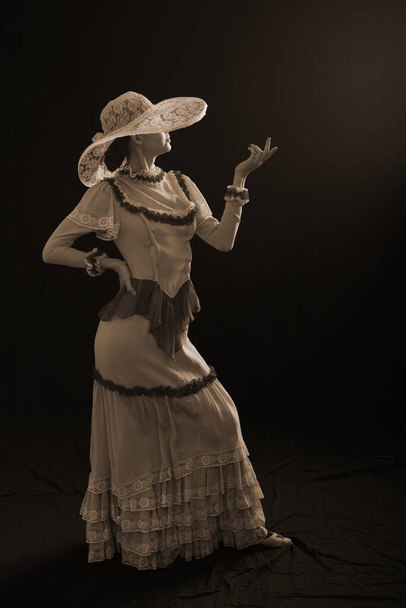A girl in an old dress and hat stands on a dark background and is sad - Zdjęcie, obraz