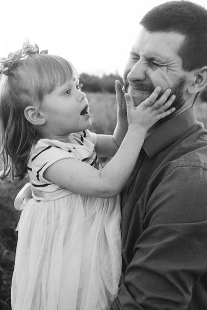 Isä ja tytär leikkivät yhdessä kukkakentällä. Onnellinen ja huolehtiva isä pitää hauskaa tyttärensä kanssa. Isä kiinnittää huomiota lapseensa. Parrakas mies ja pieni tyttö kukkia hiuksissaan - Valokuva, kuva