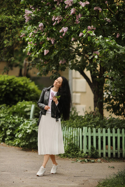 Young model woman in white skirt and black leather jacket is enjoying blooming lilac tree on the spring day - Φωτογραφία, εικόνα