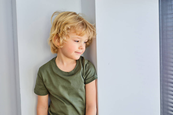 Adorable little blond kid boy standing near window and looking at side, indoors - Foto, imagen