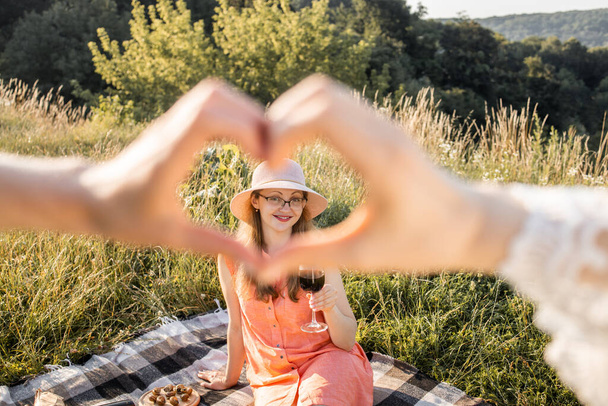 View through the heart sign of pretty woman in dress and hat, sitting on checkered blanket outdoors - Fotó, kép