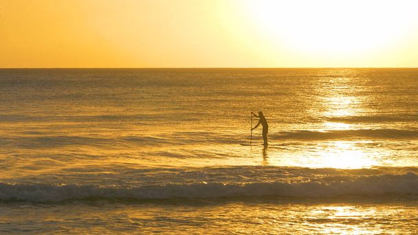 SILHOUETTE: Un hombre irreconocible se levanta en los paddleboards en una soleada noche de verano. - Foto, imagen