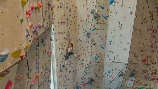 AERIAL: Climber girl climbs up an indoor climbing route filled with jug holds. - Photo, Image