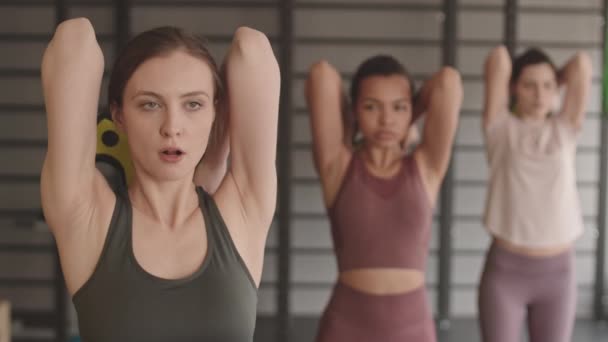 Vyötärö-up hoikka valkoihoinen nainen tekee triceps laajennuksia barbell paino sekä hämärtynyt urheilijat seisoo hänen takanaan kuntosalilla - Materiaali, video