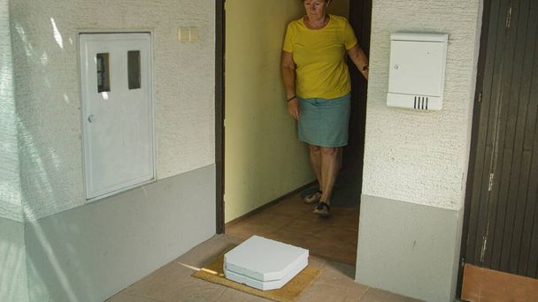 Starší žena otevře přední dveře vyzvednout dvě krabice od pizzy vlevo na jejím prahu. - Fotografie, Obrázek