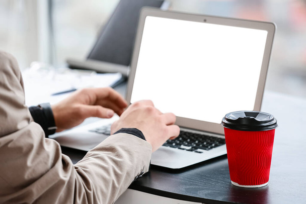 Νεαρός με φλιτζάνι καφέ χρησιμοποιώντας φορητό υπολογιστή στο γραφείο - Φωτογραφία, εικόνα