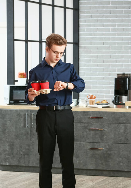 Giovane uomo con tazze di caffè guardando orologio da polso in ufficio - Foto, immagini