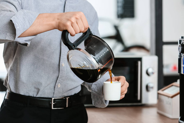 Young man pouring coffee into cup in office, closeup - 写真・画像