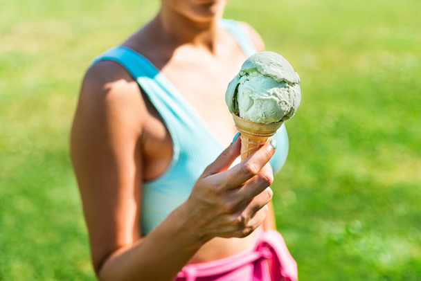 Παγωτό χωνάκι στο χέρι μιας γυμνασμένης γυναίκας το καλοκαίρι. Ζελατίνα και πολύχρωμη μόδα στο πάρκο. Retro vibes in outfit and icecream on sunny week. Τρώγοντας και απολαμβάνοντας καλοκαιρινές διακοπές έξω. Πράσινο γρασίδι. - Φωτογραφία, εικόνα