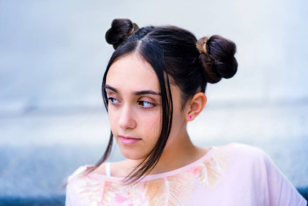 Pensive stylish teenage brunette female with hair buns looking away dreamily against blurred background - Photo, Image