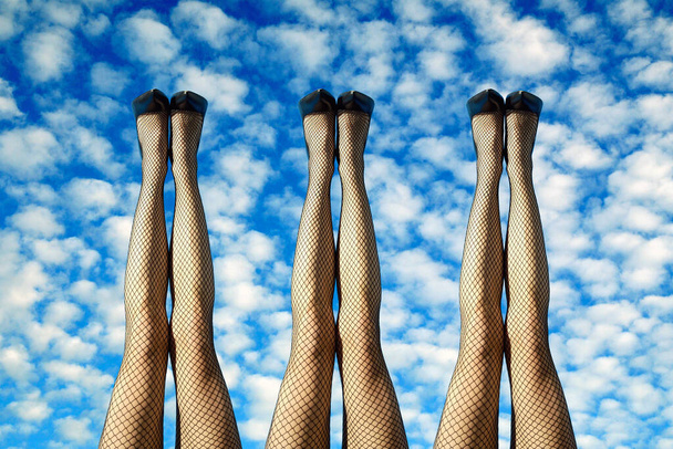 3 pairs of sexy female legs in black fishnet stockings and stilettos raised up side by side against a cloudy sunny blue sky in a conceptual image - Fotoğraf, Görsel