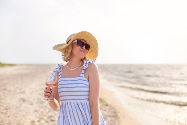 Χαρούμενη ενήλικη γυναίκα με καλοκαιρινά ρούχα χαμογελώντας και κοιτάζοντας μακριά απολαμβάνοντας το κρασί στην αμμώδη παραλία κοντά στη θάλασσα - Φωτογραφία, εικόνα