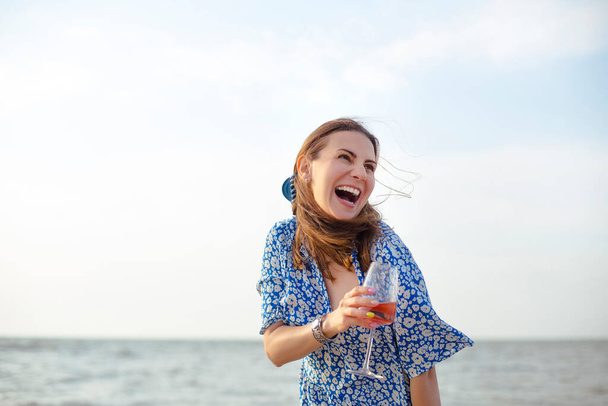 Deniz kıyısındaki kumlu sahilde şarap içerken gülümseyen ve bakışlarını çeviren mutlu yetişkin kadın. - Fotoğraf, Görsel