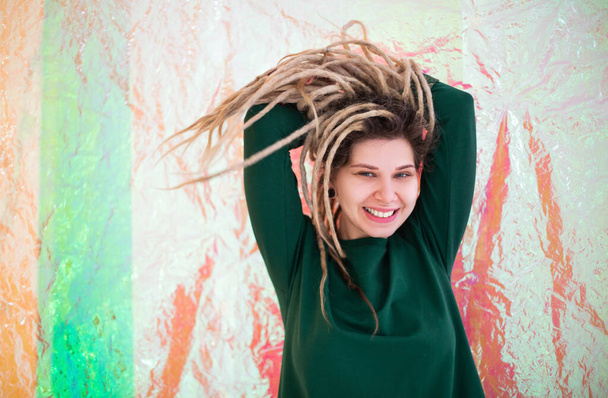 Veselá mladá žena v pestré oblečení při pohledu na kameru s úsměvem a třesoucí dredy izolované na holografickém pozadí - Fotografie, Obrázek