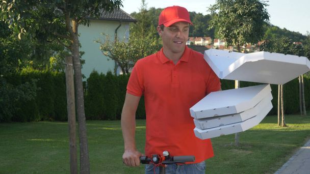 CLOSE UP: Courier leveren pizza 's druppels een kartonnen doos tijdens het rijden e-scooter - Foto, afbeelding