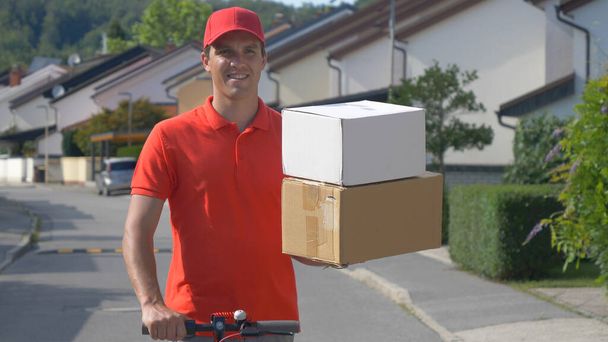 CERRAR: El repartidor de paquetes sostiene dos cajas en una mano mientras monta el scooter. - Foto, imagen