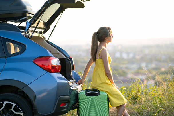 Young woman resting on a green suitcase near her car in summer nature. Travel and vacations concept. - 写真・画像