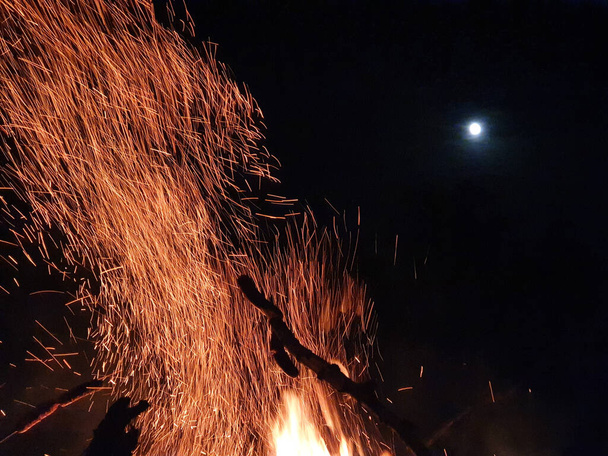 Близко: Светящиеся оранжевые искры вылетают из пылающего огня в ночное небо. - Фото, изображение