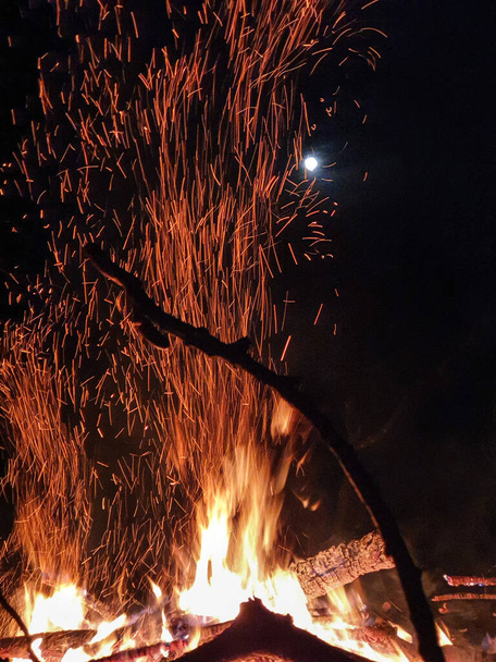 Светящиеся оранжевые искры вылетают из пылающего огня в ночное небо. - Фото, изображение