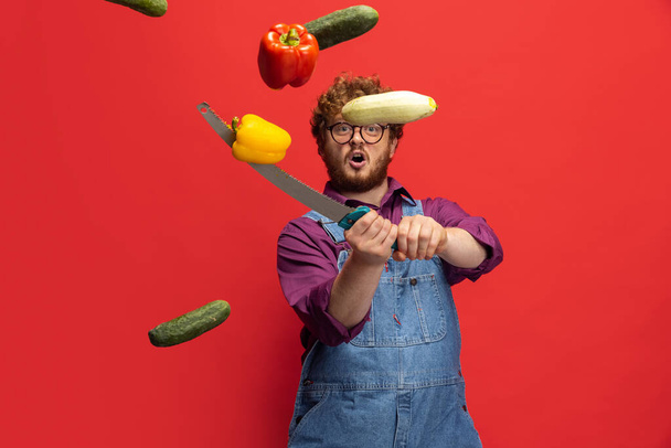 Flying vegetables. Cute redheaded bearded man, farmer with knife having fun isolated over red studio background. Concept of professional occupation, work. - Valokuva, kuva