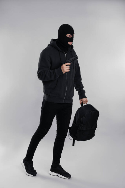 Man wearing black balaclava with backpack and gun on light grey background - Foto, imagen