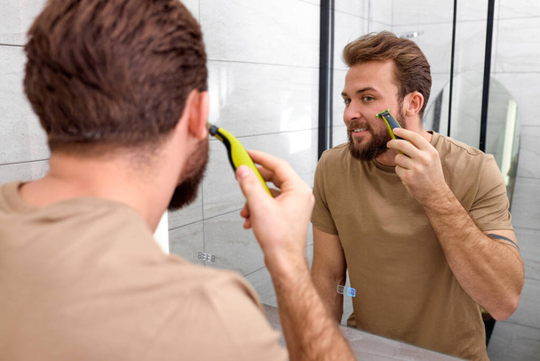 beauty, shaving. man looking at mirror, shaving beard with electric shaver - Foto, imagen