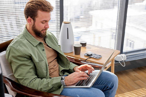 Молодий білий чоловік працює тримаючи ноутбук, дивлячись на екран комп'ютера, онлайн-навчання
 - Фото, зображення