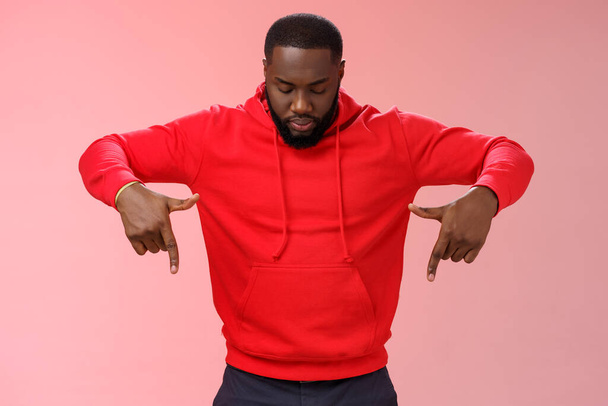 Focused interested serious-looking young african american bearded strong guy wearing red hoodie bend head look pointing down curiously, intrigued what happening lower floor, standing pink background - Photo, Image