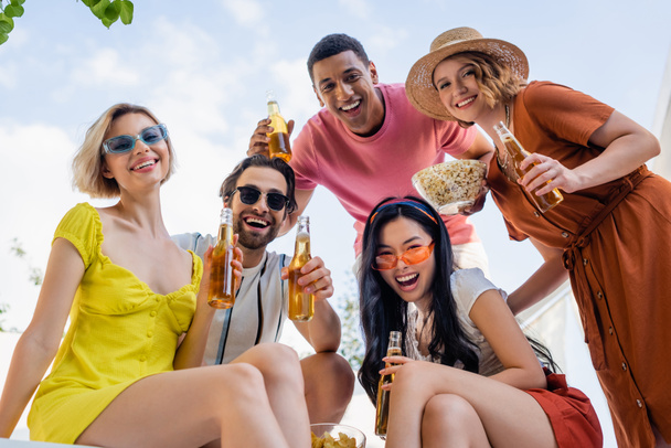 low angle view of multiethnic friends with beer and snacks smiling at camera outdoors - Photo, Image