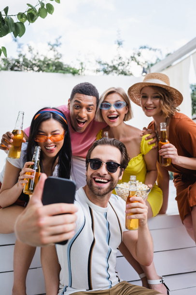 Güneş gözlüklü mutlu adam verandada biralı çok ırklı arkadaşlarıyla selfie çekiyor. - Fotoğraf, Görsel