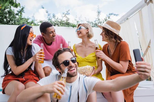 Fröhlicher Mann mit Sonnenbrille macht Selfie in der Nähe von Freunden, die Bier trinken - Foto, Bild