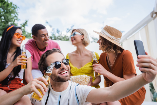 uomo gioioso scattare selfie su smartphone durante la festa estiva con amici multietnici offuscati - Foto, immagini