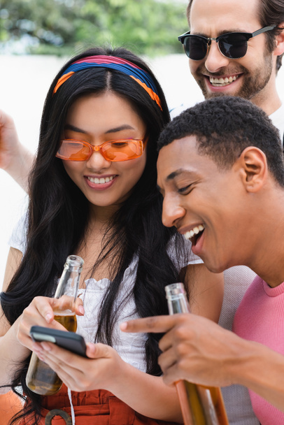 excited multiethnic friends looking at mobile phone during beer party - Photo, Image