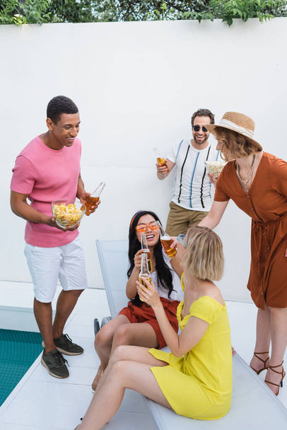 glückliche interrassische Frauen, die mit Bier anstoßen, während sie auf der Terrasse neben glücklichen Freunden sitzen - Foto, Bild