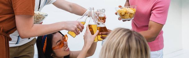 многонациональные друзья звон бутылки пива возле чипсов и попкорна во время вечеринки, баннер - Фото, изображение